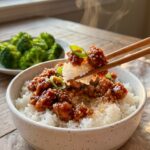 A close-up of chopsticks lifting a bite of sticky firecracker chicken and white rice from a bowl.
