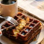 A close-up of a knife and fork cutting into a crispy French toast waffle, showing the soft, fluffy interior with steam rising.
