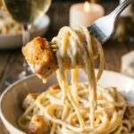 A close-up of a fork holding a piece of seared garlic butter chicken and twirled creamy pasta, with sauce dripping slightly