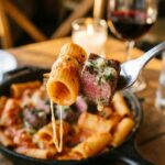 A close-up of a fork holding a creamy rigatoni noodle and a juicy steak tip, with a slight cheese pull.