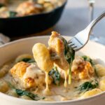 A close-up of a fork lifting a piece of creamy chicken and a pillowy gnocchi from a bowl, with white wine in the background.