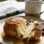 A close-up of a fork cutting into a fluffy piece of Hawaiian roll French toast drenched in maple syrup, with a coffee mug in the background.