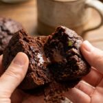 A close-up of a chocolate zucchini muffin broken in half, showing a moist, fudgy center with melted chocolate.