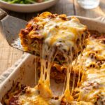 A close-up of a spatula lifting a heavy, cheesy square of beef and noodle casserole with a long cheese pull stretching to the pan.