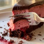 A close-up of a fork cutting through a slice of layered raspberry chocolate mousse cake, showing the soft textures.