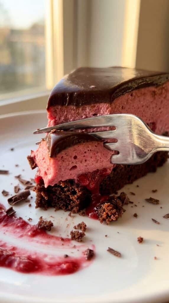 A close-up of a fork cutting through a slice of layered raspberry chocolate mousse cake, showing the soft textures.