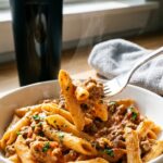 A close-up of a fork holding a bite of creamy beef pasta, with a protein shaker bottle blurred in the background.