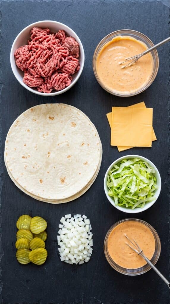 A flat lay showing ground beef, tortillas, American cheese, shredded lettuce, pickles, onions, and secret sauce on a dark slate board.