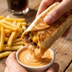 A close-up of a hand dipping a crispy beef and cheese wrap into a small bowl of creamy orange burger sauce, with fries in the background.