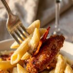 A close-up of a fork holding creamy penne pasta, a piece of honey pepper chicken, and bacon.