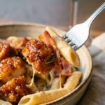 A close-up of a fork lifting a bite of creamy pasta, glazed chicken, and bacon from a bowl.