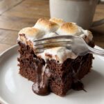 A close-up of a fork cutting into a slice of chocolate poke cake, showing gooey hot fudge oozing from the center, with a mug of hot cocoa in the background.