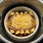 A view inside an Instant Pot stainless steel liner showing a ceramic casserole dish filled with uncooked cheesy enchiladas sitting on a metal trivet.