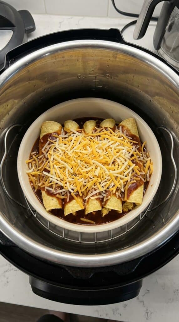 A view inside an Instant Pot stainless steel liner showing a ceramic casserole dish filled with uncooked cheesy enchiladas sitting on a metal trivet.