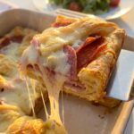 A spatula lifting a slice of Italian crescent bake, showing a cheese pull and layers of deli meat, with a side salad in the background.