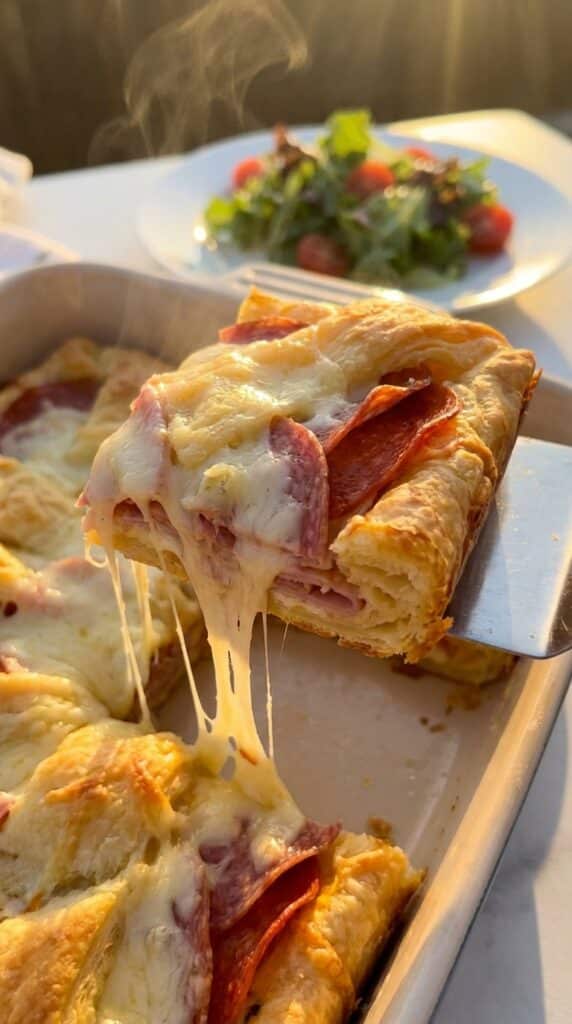 A spatula lifting a slice of Italian crescent bake, showing a cheese pull and layers of deli meat, with a side salad in the background.