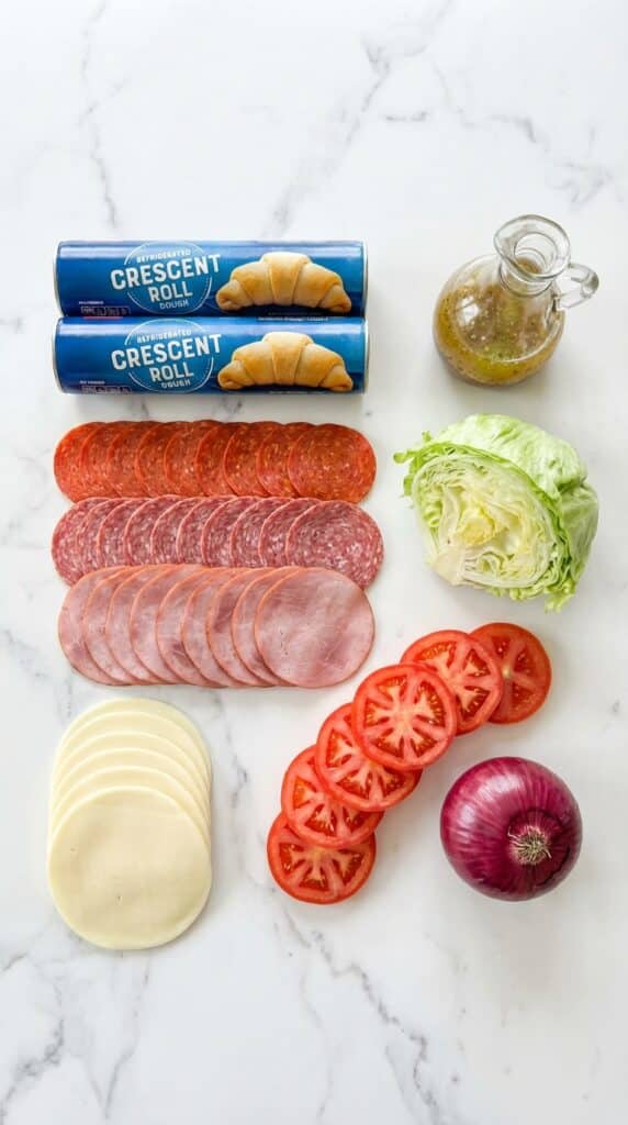 A flat lay showing crescent dough tubes, deli meats (salami, ham, pepperoni), provolone cheese, lettuce, tomatoes, and Italian dressing on a marble counter.