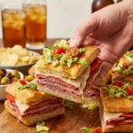A close-up of a hand lifting an Italian sub square, showing layers of meat, melted cheese, and fresh salad topping.