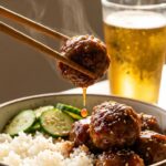 A close-up of chopsticks lifting a glossy, sauce-covered meatball from a rice bowl with cucumbers.