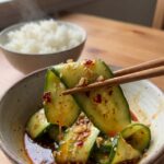 A close-up of wooden chopsticks lifting a glossy, seasoned slice of cucumber salad with a bowl of rice in the background.