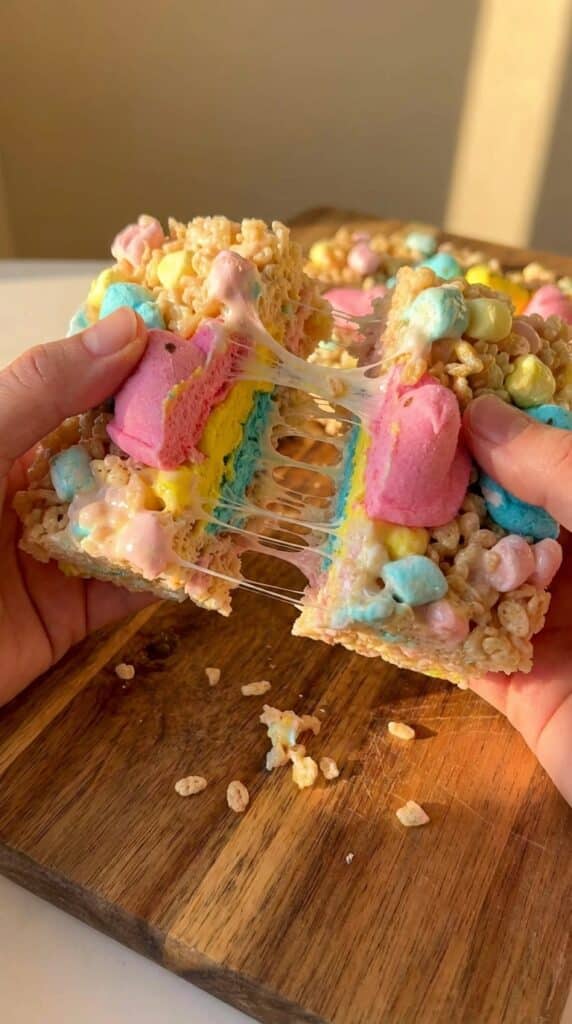 Two hands pulling apart a colorful layered rice krispie treat, showing a gooey marshmallow stretch.