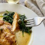 A close-up of a fork cutting into a piece of lemon chicken scallopini over wilted spinach on a white plate.