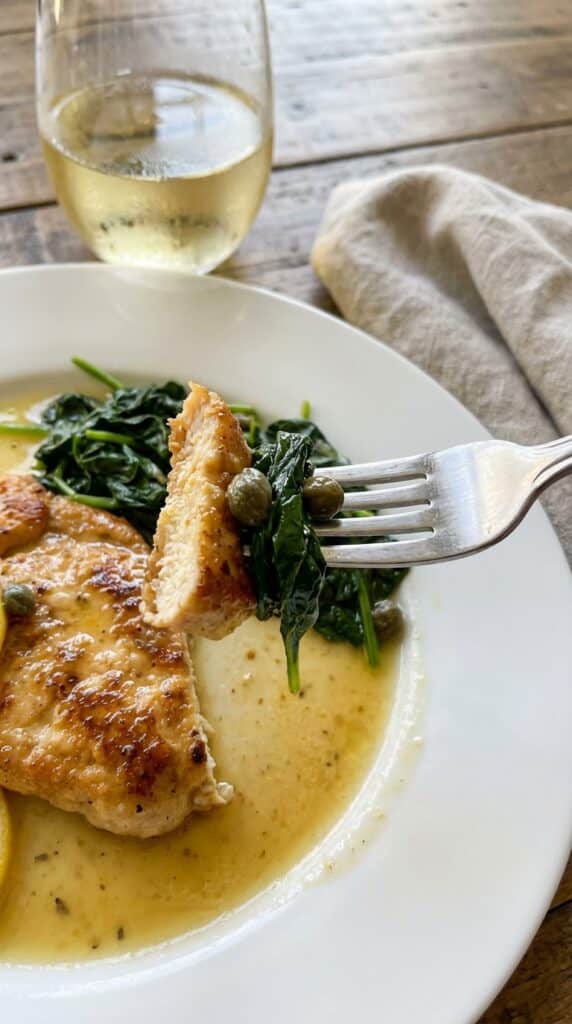 A close-up of a fork cutting into a piece of lemon chicken scallopini over wilted spinach on a white plate.