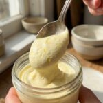 A close-up of a silver spoon lifting a thick, smooth dollop of lemon mousse with specks of zest visible.