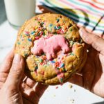 Two hands holding a massive, thick baked cookie topped with a pink frosted animal cookie and rainbow sprinkles, with a glass of milk in the background.
