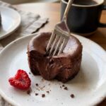 A close-up of a fork cutting into a creamy mini chocolate cheesecake on a white plate with a cup of coffee in the background.