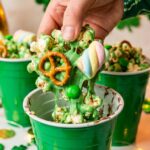 A close-up of a hand holding a cluster of green chocolate-covered popcorn, pretzels, and marshmallows taken from a party cup.