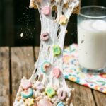 Two hands breaking a Lucky Charms marshmallow treat in half, showing a long, gooey marshmallow stretch.
