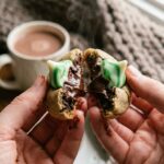 Hands breaking a warm chocolate mint blossom cookie in half, showing a soft, fudgy center and a melting mint kiss.