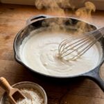 A close-up of a whisk stirring a thick, bubbling creamy white sauce in a skillet with dry powder mix nearby.