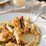 A close-up of a fork holding a perfect bite of creamy chicken pasta with a sun-dried tomato, with wine and candles in the background.