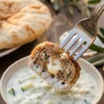 A close-up of a fork holding a halved chicken meatball with visible feta inside, dipping it into creamy tzatziki sauce.