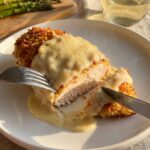A close-up of a knife and fork cutting into a juicy chicken breast coated in a creamy, golden Caesar sauce, with asparagus in the background.