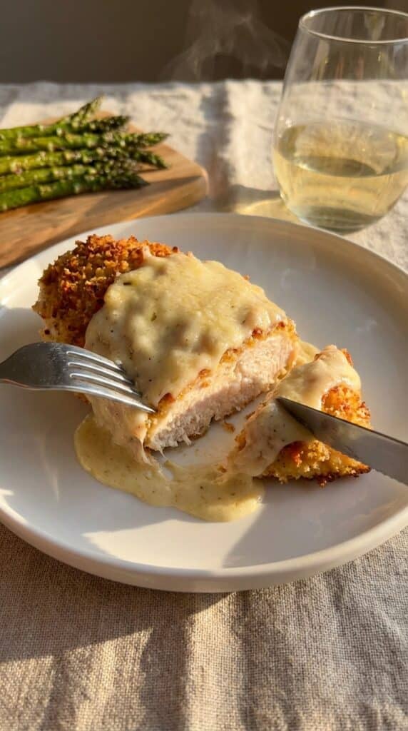 A close-up of a knife and fork cutting into a juicy chicken breast coated in a creamy, golden Caesar sauce, with asparagus in the background.