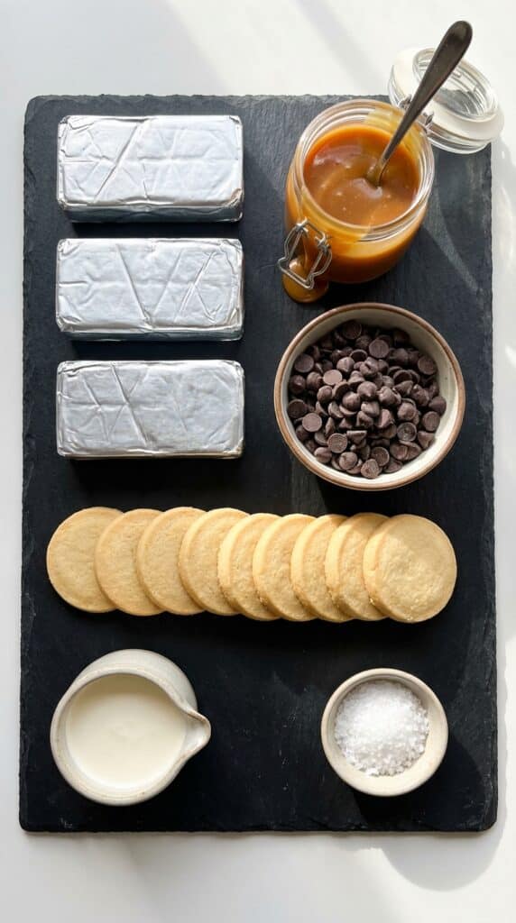 A flat lay showing cream cheese, caramel sauce, chocolate chips, shortbread cookies, and flaky sea salt on a dark slate board.