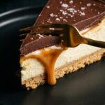 A close-up of a slice of millionaire cheesecake on a black plate, with a gold fork cutting into it, showing the layers and a caramel drip.