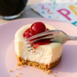 A close-up of a dessert fork cutting into an unwrapped mini cherry cheesecake on a pink plate, showing the creamy texture.
