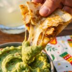 A close-up of a hand dipping a cheesy, crispy mini chicken quesadilla into a bowl of guacamole, with a visible cheese pull.