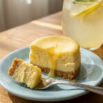 A close-up of a mini lemon cheesecake with a bite taken out by a fork, showing the creamy texture and crumbly crust.