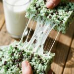 A close-up of hands pulling apart a green mint chocolate chip Rice Krispie treat, showing a long, gooey marshmallow stretch.