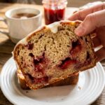 A close-up of a hand holding a thick slice of strawberry bread, showing the moist texture and jammy fruit pockets.