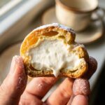 A close-up of a hand holding a bitten cream puff, showing the hollow pastry shell completely filled with white cream.