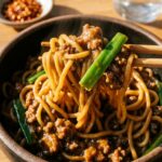 A close-up of wooden chopsticks lifting saucy Mongolian beef noodles out of a dark bowl.