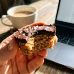 A close-up of a hand holding a peanut butter oat cup with a bite taken out, with a coffee mug in the blurred background.