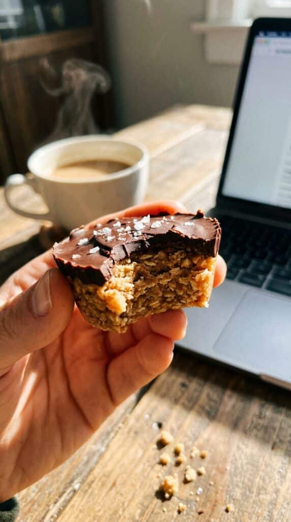 A close-up of a hand holding a peanut butter oat cup with a bite taken out, with a coffee mug in the blurred background.