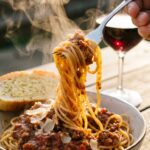 A close-up of a fork twirling spaghetti coated in thick meat sauce with a piece of garlic bread in the background.